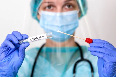 Eine Frau in medizinischer Schutzkleidung h&auml;lt vor ihrem K&ouml;rper ein Testst&auml;bchen und ein Testr&ouml;hrchen, das mit COVID-19 Coranvirus beschriftet ist