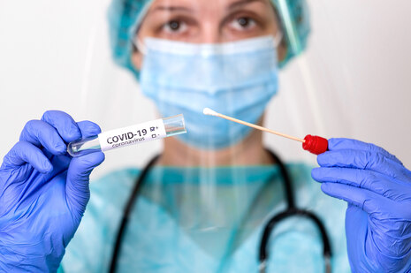 Eine Frau in medizinischer Schutzkleidung hält vor ihrem Körper ein Teststäbchen und ein Teströhrchen, das mit COVID-19 Coranvirus beschriftet ist