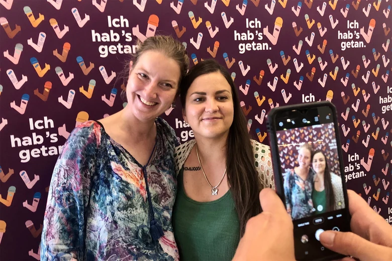 Zwei jungen Frau werden von einer dritten Person mit dem Handy vor der "Ich hab's getan"- Selfie-Wand im Impfzentrum fotografiert