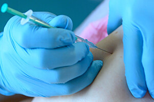 Jemand mit blauen medizinischen Handschuhen sticht eine Spritze mit dem Anti-Corona-Impfstoff in den Oberarm einer Person. Es handelt sich um eine Detailaufnahme des Impfvorganges.